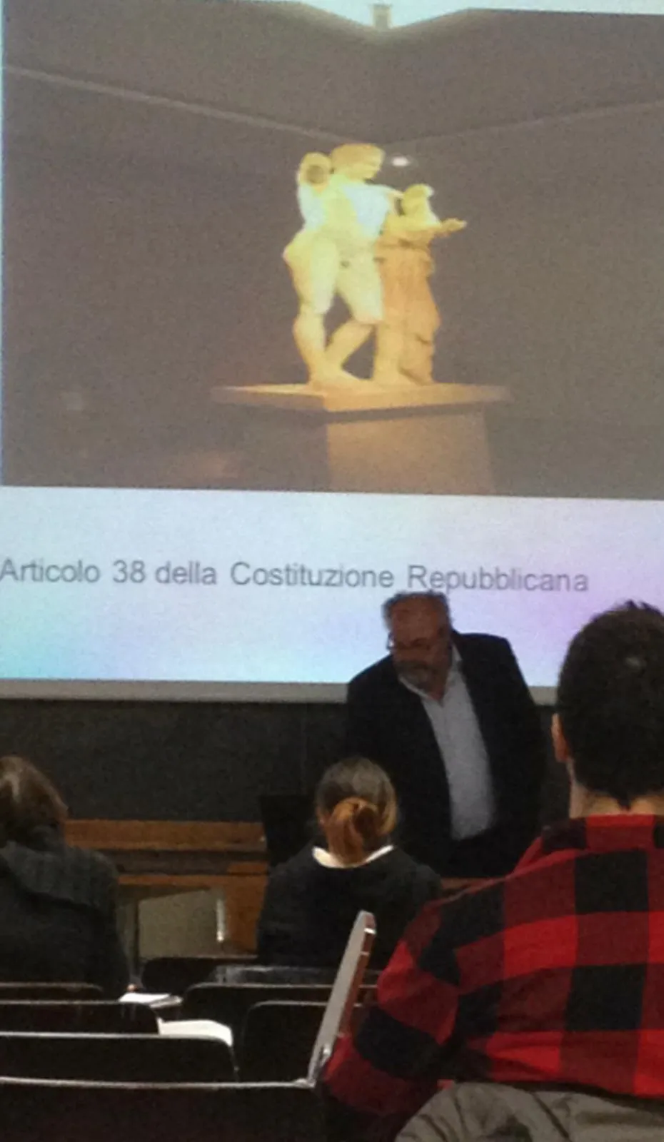 Un relatore davanti alla proiezione di una statua classica con la didascalia «Articolo 38 della Costituzione Repubblicana».