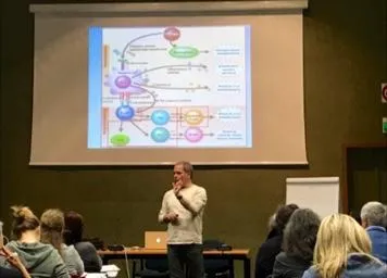 Un relatore davanti a uno schema di vie biologiche proiettato, mentre il pubblico prende appunti.