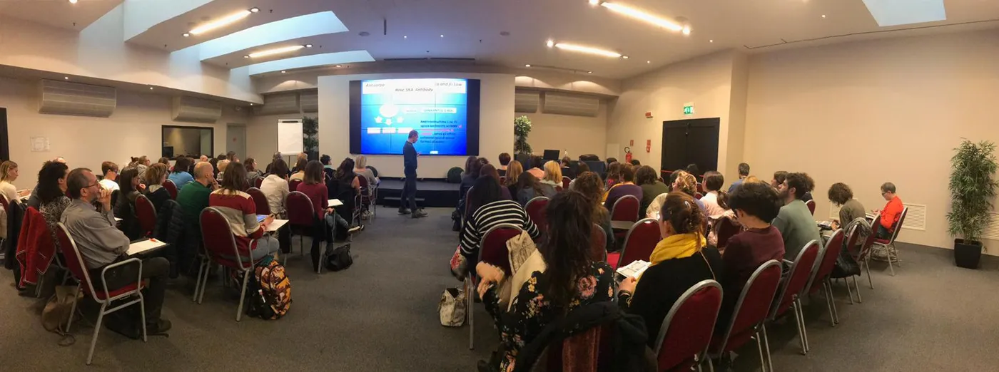 Panoramica di una sala convegni piena: file di sedie rosse, pubblico di spalle e un relatore davanti a un grande schermo.