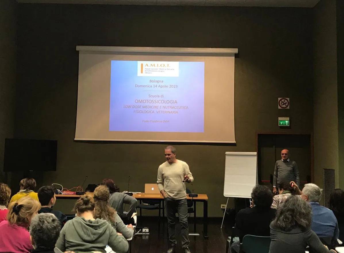 Un relatore parla davanti allo schermo con la slide di una scuola di omotossicologia; un secondo relatore attende sul fondo della sala.