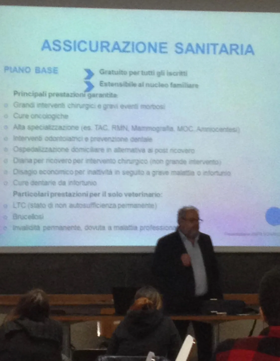 Un relatore in piedi accanto alla slide «Assicurazione sanitaria», con l’elenco delle prestazioni garantite.