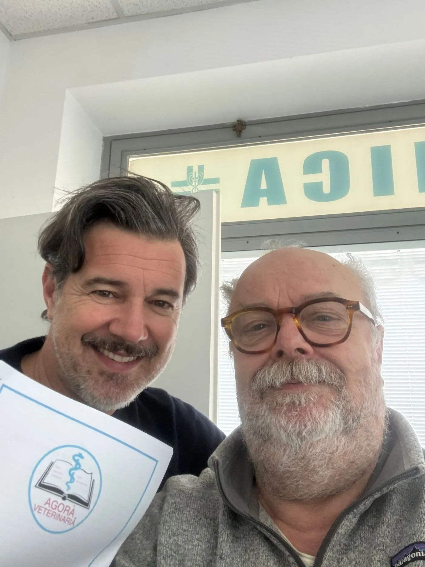 Due uomini sorridenti in posa davanti a una finestra; uno dei due tiene in mano il manifesto con il logo di Agorà Veterinaria.